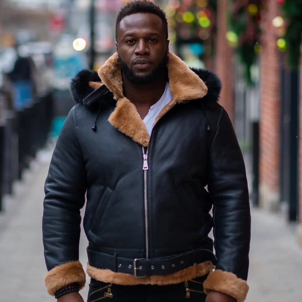 Front view of a man wearing Jean's B3 Bomber Aviator Shearling Jacket with hoodie, featuring sleek black leather with contrasting tan shearling lining around the collar and cuffs. The jacket includes a front zipper closure and leather belt detailing at the waist, perfect for colder weather.