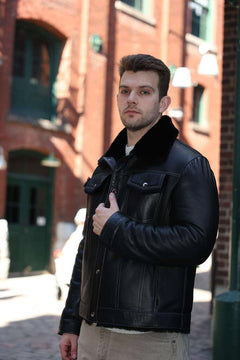 Man wearing a black leather jacket standing in an urban setting with brick buildings.