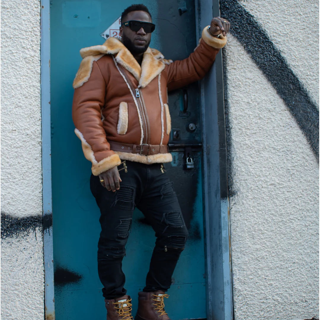 Alek's Biker Shearling Jacket with Fur Details, worn by a man standing and leaning against a door with one arm raised, highlighting the fur collar, waist belt, and brown leather finish. The jacket offers a stylish and rugged look with plush fur accents.