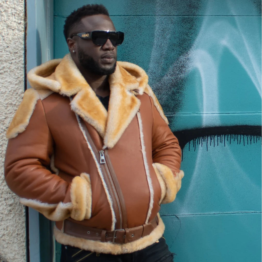 Side view of Alek's Biker Shearling Jacket with Fur Details, worn by a man, showcasing the unique contrast of the brown leather jacket with the beige fur-lined collar and cuffs. The model poses in front of a teal-colored wall.