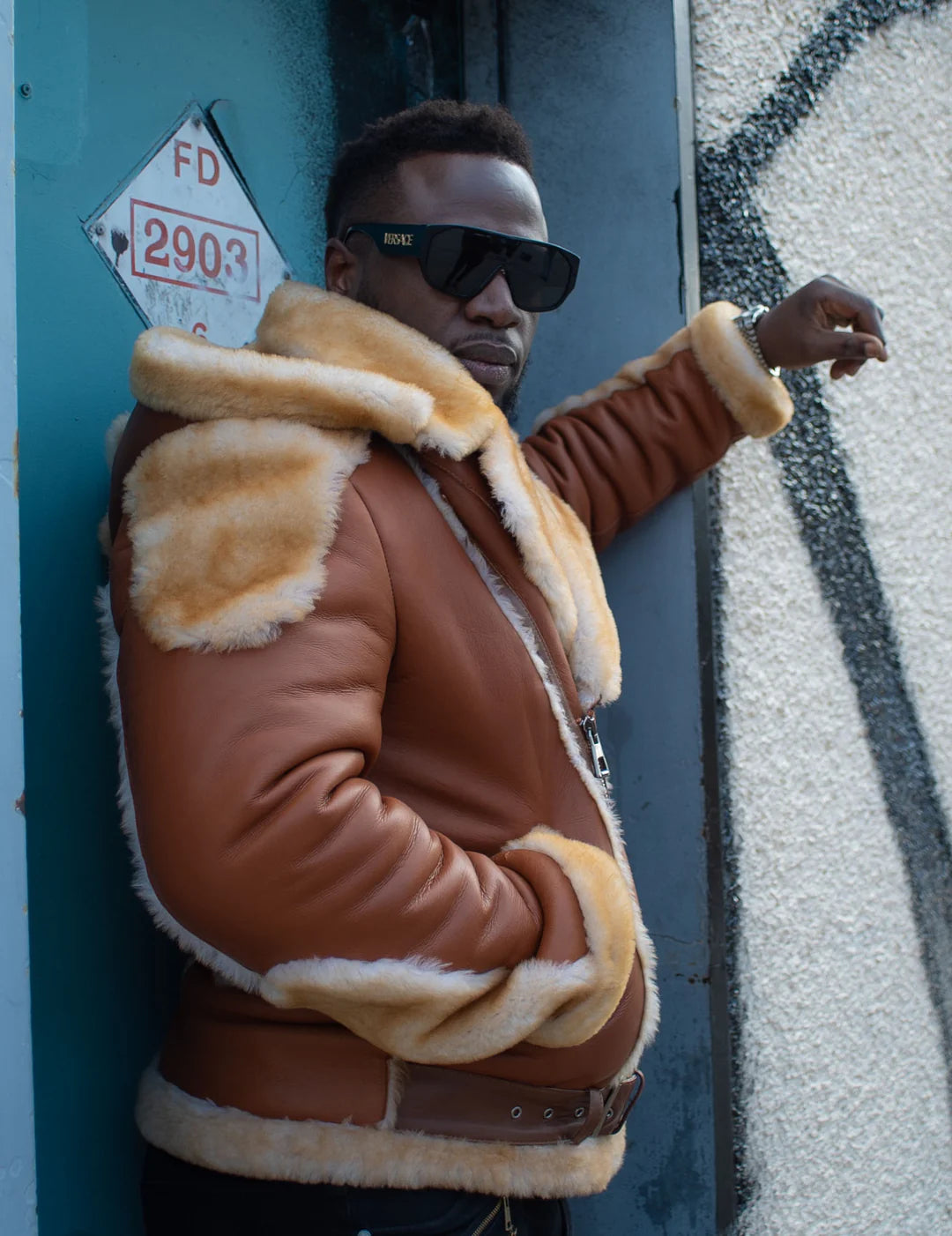 Alek's Biker Shearling Jacket with Fur Details, shown worn by a man standing against a blue door, featuring a zipper front and fur-lined collar and cuffs. The jacket is styled with a brown leather body and beige fur accents.