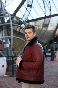 Man wearing Aurelius Burgundy B3 Bomber Jacket with black fur trim standing in front of a large metal sculpture.