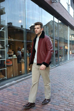 Man in Aurelius Burgundy B3 Bomber Jacket walking on a brick sidewalk with a store in the background