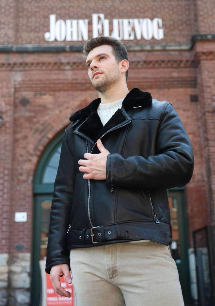 Man wearing a black leather jacket with fur collar in front of a brick building with 'John Fluevog' sign.