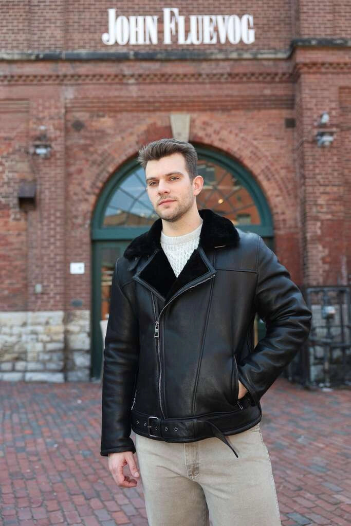 Man wearing a black leather jacket with fur collar in front of a brick building with 'John Fluevog' sign.