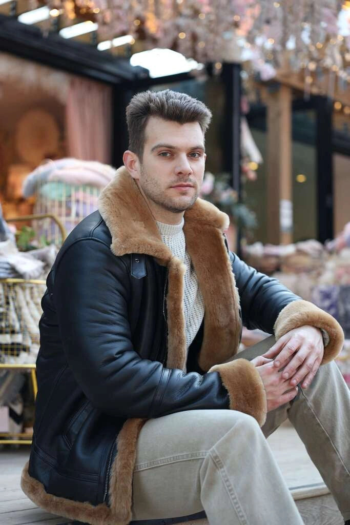 Man wearing a dark Classic Ginger Brown B3 Bomber Aviator Shearling Jacket with fur trim sitting outdoors.
