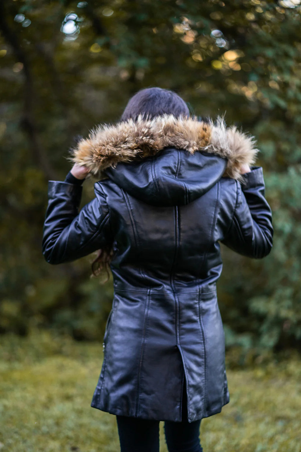 Woman back profile in Jazmyn's long winter coat, emphasizing the thick fox fur hood and the protective longline fit in a forest setting.