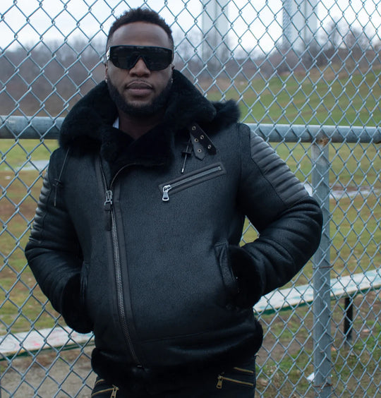 Man wearing a black aviator shearling bomber jacket and sunglasses standing in front of a chain-link fence.