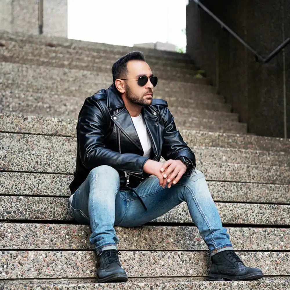 A male model wearing sunglasses and the Perfect Biker Style heavy leather biker jacket, sitting on concrete steps that highlights the asymmetrical zipper and belted waist.