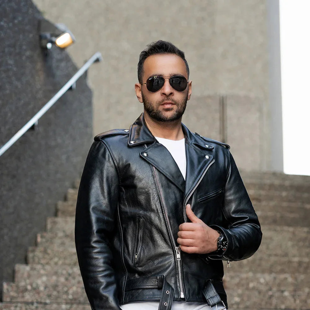 A male model wearing the Perfect Biker Style heavy leather biker jacket, standing in a classic leaning pose that highlights the asymmetrical zipper and belted waist.