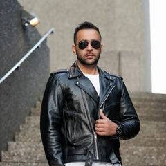 A male model wearing the Perfect Biker Style heavy leather biker jacket, standing in a classic leaning pose that highlights the asymmetrical zipper and belted waist.