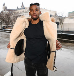 Lifestyle image of a model wearing Ridgeway's Vintage Distressed Brown Shearling Vest with a cozy shearling-lined hoodie. The rugged brown leather vest features soft beige faux fur lining, with the model standing outdoors in a stylish urban setting.